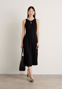 Abito nero senza maniche con scollatura a chiave, silhouette fluida e lunghezza fino al polpaccio, abbinato a sandali neri con cinturini e una grande borsa a tracolla.