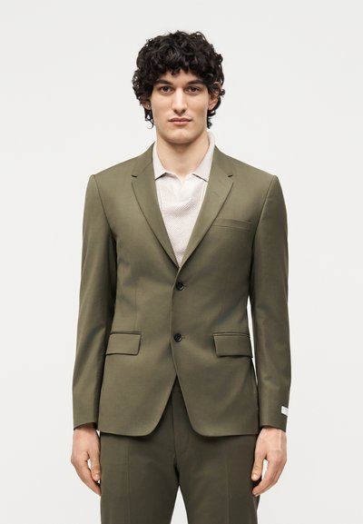 Νέος άνδρας με σγουρά μαλλιά που φοράει ένα ελιά χρώματος κοστούμι και ένα λευκό υφασμάτινο πουκάμισο, στέκεται μπροστά σε ένα απλό φωτεινό φόντο.