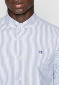 Koszula na guziki z pionowymi niebiesko-białymi paskami. Posiada ostro zakołnierz i małe haftowane logo po lewej stronie. Materiał bawełniany.