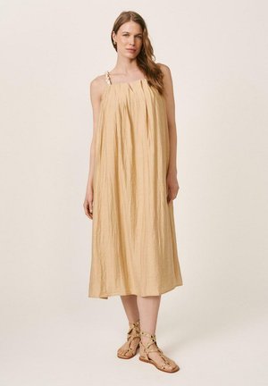 Mujer con un vestido midi beige suelto sin mangas, con tirantes trenzados y sandalias planas con tachuelas, de pie contra un fondo claro y sencillo.