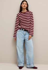 Maglione a righe bordeaux e bianco con collo rotondo, abbinato a jeans a gamba larga azzurri e scarpe rosso scuro, su uno sfondo neutro.