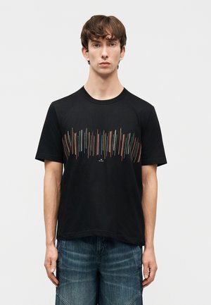 Νεαρός άντρας που φοράει μαύρο μπλουζάκι με πολύχρωμες κάθετες γραμμές στο στήθος και μπλε τζιν, στέκεται μπροστά σε απλό φόντο.