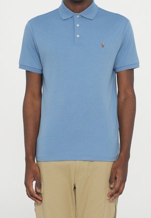 Mand iført lyseblå kortærmet polo med tre knapper og beige bukser stående mod en ensfarvet baggrund.
