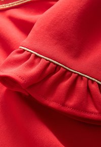 Detalje af rødt tøj med en flæsekanter med guldkant, glat tekstur og ensfarvet design.