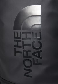 Svart ryggsekk med en strukturert overflate og en fremtredende blank logo i mørkegrått. Designet er strømlinjeformet med fet skrift.