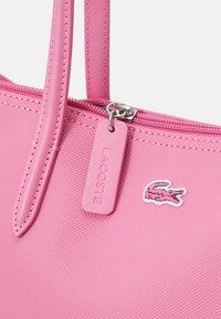 Sac à main synthétique rose avec une surface texturée, doté d'une fermeture éclair, de deux poignées et d'une étiquette logo Lacoste. Design minimaliste.