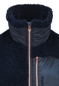 Marinblå fleecejacka med hög krage, med en kviltad tygdel, två fickor med dragkedja och detaljer i roséguld.