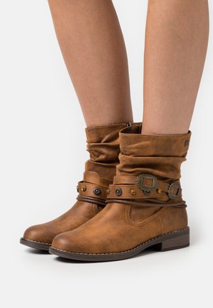 Bruine slouchlaarzen tot halverwege de kuit met decoratieve riemen, metalen studs en gespen, gedragen op blote benen tegen een witte achtergrond.