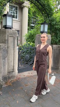 Top fără mâneci din in maro și pantaloni asortați, purtând adidași albi. Ținând o udătoare albă, stând lângă o poartă decorativă.