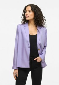 Lavendel blazer van gladde stof, met een enkele knoopsluiting en gelaserde revers, gecombineerd met een zwarte top en strakke zwarte broek.