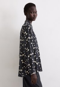 Vrouw in een losse zwarte knoopshirt met een wit abstract bloempatroon, staande zijwaarts tegen een eenvoudige witte achtergrond.