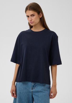 Frau mit glattem braunem Haar trägt ein lockeres dunkelblaues T-Shirt und blaue Jeans, steht vor einem schlichten, hellgrauen Hintergrund.
