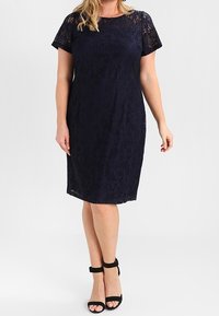Abito in pizzo blu navy con maniche corte e taglio aderente, caratterizzato da un motivo floreale e un orlo al ginocchio. Abbinato a sandali neri con cinturini.