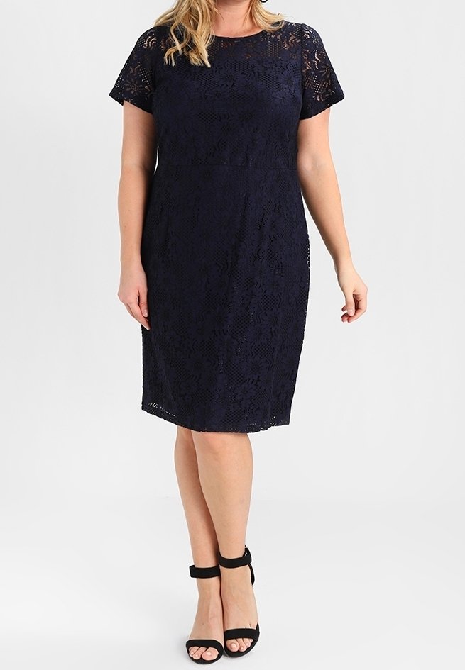 Abito in pizzo blu navy con maniche corte e taglio aderente, caratterizzato da un motivo floreale e un orlo al ginocchio. Abbinato a sandali neri con cinturini.