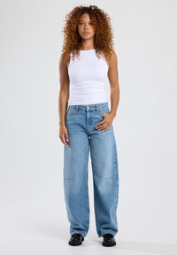 Witte mouwloze top gecombineerd met losse lichtblauwe jeans. De jeans hebben een recht model en een lichte vervaging, met zichtbare stikseldetails.
