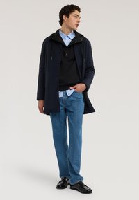 Marineblauer wasserdichter Mantel mit Kapuze, darüber ein schwarzer Pullover und ein hellblaues Hemd, kombiniert mit hellblauen Jeans und schwarzen Schuhen.