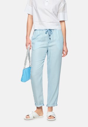 Woman wearing light blue drawstring pants, white short-sleeve shirt, white braided sandals, and holding a small blue purse with a chain strap.
