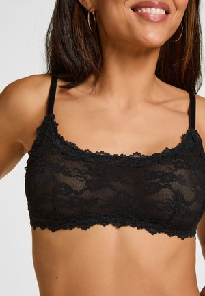 Frau trägt eine schwarze Spitzen-Bralette mit dünnen Trägern und Blumenmustern, lächelt, goldene Creolen sichtbar.