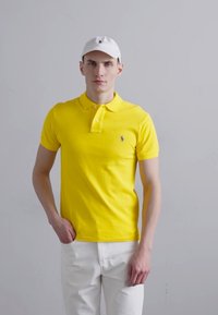 Geltona medvilninė polo marškinėlių su apykakle ir trimis mygtukais, derinami su baltais keliais. Marškiniai turi mažą logotipą ant krūtinės.