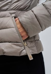 Hand die in ritszak van taupekleurig donzen jack schuift met "Salsa Jeans"-logo op de mouw en goudaccenten aan de ritstrekker.