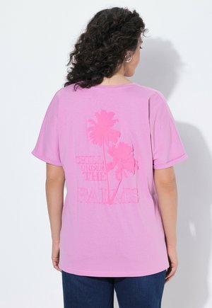 Frau mit lockigem Haar in einem pinken T-Shirt mit Palmen-Motiv und dem Schriftzug „Chill under the palms“ auf dem Rücken, trägt dunkelblaue Hose.
