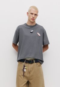 Grå kortærmet t-shirt med pink og blå grafik, parret med beige bukser og et mønstret bælte med forskellige nøgleringstilbehør.