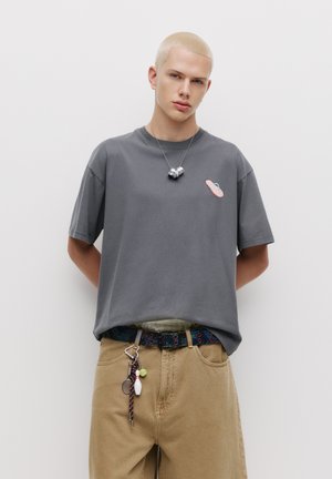 Grå kortærmet t-shirt med pink og blå grafik, parret med beige bukser og et mønstret bælte med forskellige nøgleringstilbehør.