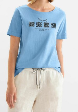 Niebieski t-shirt z krótkim rękawem z czarnym zwierzęcym nadrukiem w formie kwadratów i napisem "Mood Go wild for a while", noszony z jasnobeżowymi spodniami z troczkiem.