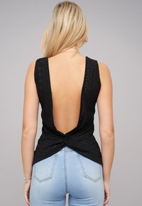 Zwarte mouwloze top met een diepe V-rug, voorzien van gestructureerde stof en een gedraaide afwerking aan de zoom. Gecombineerd met lichtblauwe jeans.