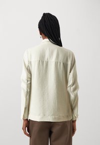 Light beige long-sleeve shirt with a textured fabric, visible seams, and a shirt collar. Worn over brown pants. Back view shown.