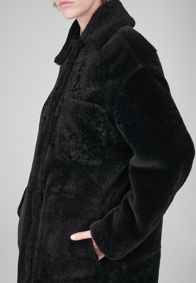 Schwarzer Kunstfellmantel mit weicher Textur, über einem Kragen, Vordertaschen und einer leicht übergroßen Passform.