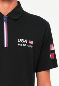 Sort polo-shirt med en lynlås krave, hvid tekst "USA SAIL GP TEAM" og et lille flag. Indeholder røde, hvide og blå accentdetaljer.