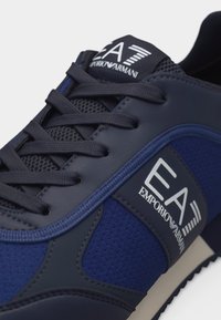 Σκούρο μπλε αθλητικό παπούτσι EA7 Emporio Armani με μαύρα κορδόνια, επάνω μέρος από δίχτυ και δέρμα, με λογότυπο στη γλώσσα και στις πλάγιες πλευρές.