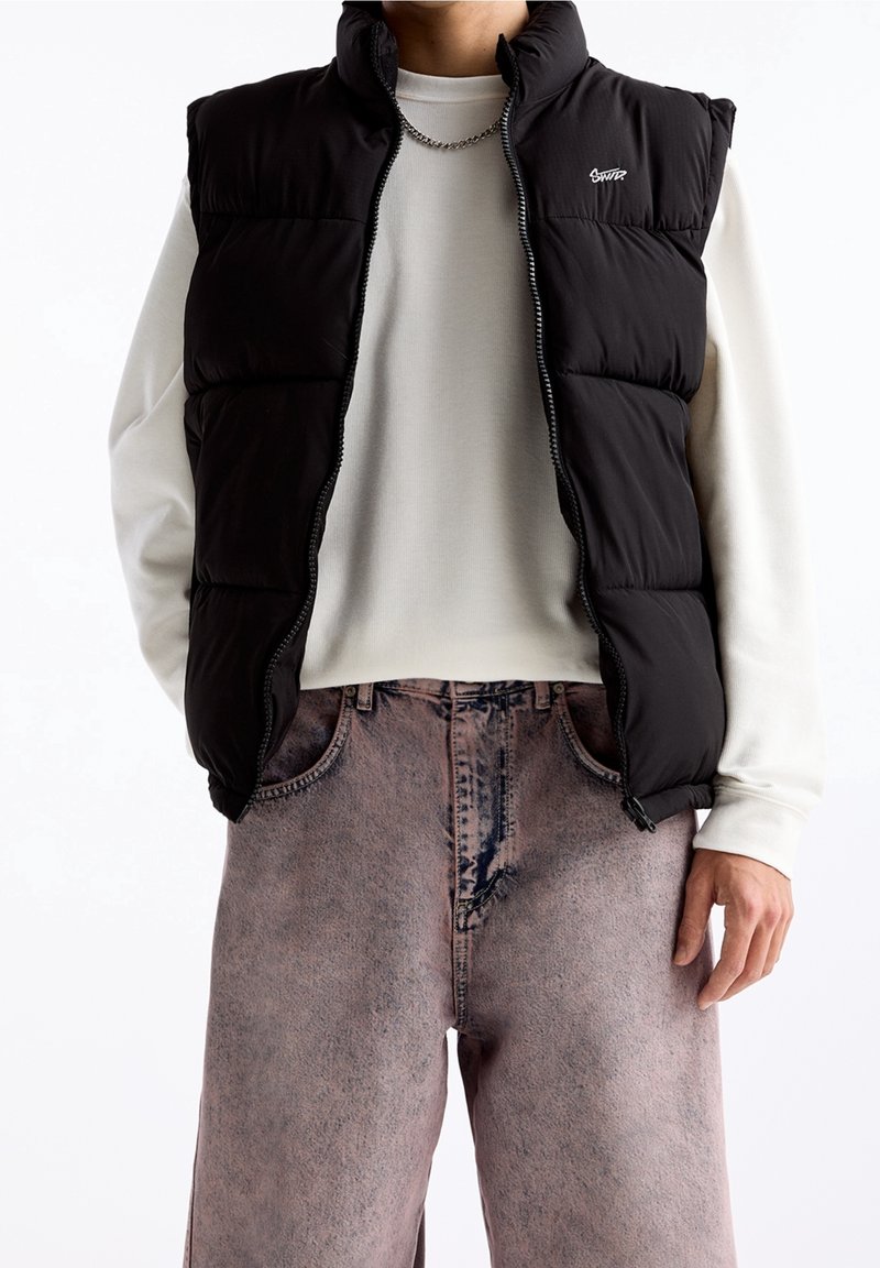 Černá prošívaná vesta s vysokým límcem a předním zipem, nošená přes světle zbarvené tričko s dlouhým rukávem a doplněná o vybledlé béžové kalhoty.