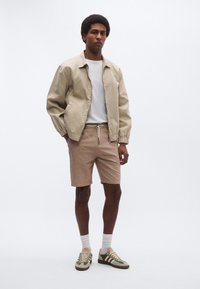 Μπεζ βομβητής μπουφάν, λευκό μπλουζάκι, ανοιχτό καφέ σορτς, συνδυασμένα με μπεζ και πράσινα αθλητικά παπούτσια. Το σύνολο διαθέτει άνετη εφαρμογή και ελαστικές λεπτομέρειες.