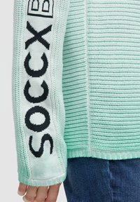 Minzgrüner, gestrickter Pullover mit gerippter Textur und weißen Ärmeln mit schwarzem "SOCCK"-Schriftzug. Gerader Saum und lässige Passform.