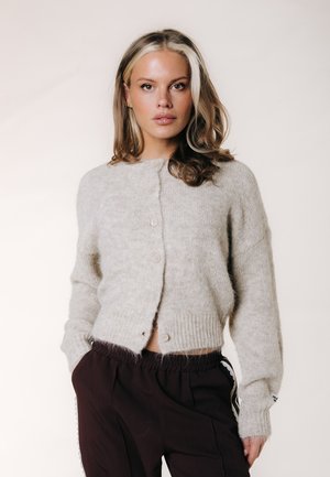 Giovane donna con capelli biondi ondulati indossa un cardigan grigio chiaro con bottoni e pantaloni scuri larghi, in piedi davanti a uno sfondo semplice.