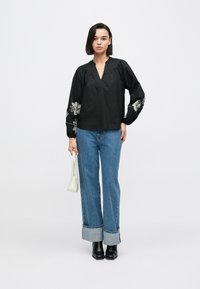 Čierna blúzka s výstrihom do „V“ a vyšívanými kvetinovými vzormi na rukávoch, kombinovaná so svetlomodrými džínsami s širokými nohavicami a čiernymi čižmami na opätku.
