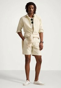 Άνδρας με κοτσίδες, φορώντας ανοιχτό μπεζ πουκάμισο με τσακισμένα μανίκια, αντίστοιχο σορτς, πλεγμένο πουλόβερ γύρω από τη μέση, λευκά sneakers και γυαλιά ηλίου.