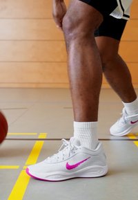 Hvide sportssko med pink detaljer og et tydeligt Nike swoosh, båret med hvide sokker, på et glat gyms gulv med bane linjer.