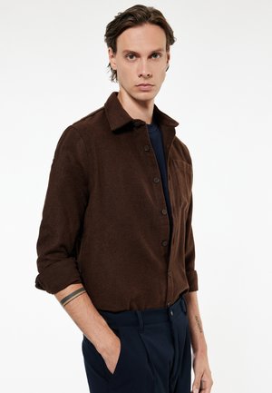 Joven con piel clara y cabello castaño, vistiendo una camisa marrón oscuro abotonada con las mangas remangadas y pantalones azul marino, de pie con una mano en el bolsillo.