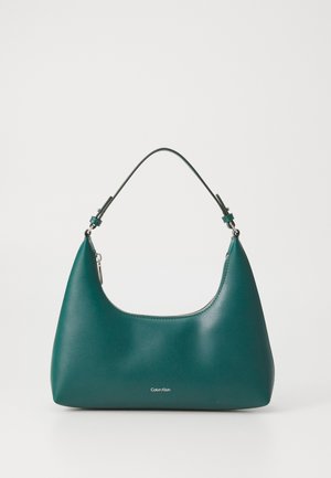 Sac à main teal en forme arrondie avec une bandoulière, arborant un logo Calvin Klein discret à l'avant sur fond blanc.