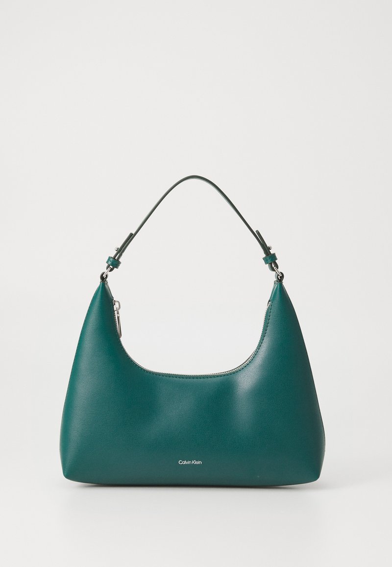 Bolso de mano en color teal con forma curva y una correa para el hombro, presentando un sutil logo de Calvin Klein en la parte frontal sobre un fondo blanco liso.