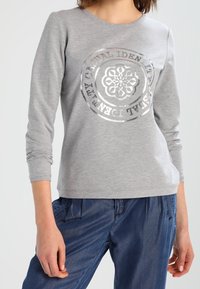 Felpa grigia con maniche lunghe, caratterizzata da un design di logo circolare in argento metallico sul davanti. Il materiale sembra morbido e strutturato.