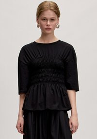 Zwarte top van zachte stof met een gathered taille, korte mouwen en een vloeiende peplum ontwerp. Gladde textuur met minimale details.