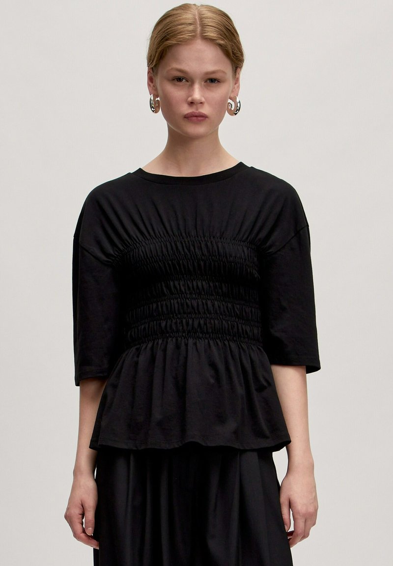 Zwarte top van zachte stof met een gathered taille, korte mouwen en een vloeiende peplum ontwerp. Gladde textuur met minimale details.