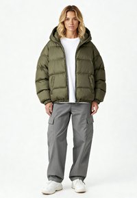 Giacca imbottita verde oliva con cappuccio, texture trapuntata, chiusura a zip e polsini elasticizzati; abbinata a pantaloni cargo grigi e sneakers bianche.