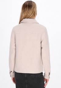 Light beige jacket with a collar, textured fabric, long sleeves, and button detail at the cuffs. Features a straight cut and seam design.