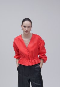 Femme à cheveux foncés, portant un chemisier rouge boutonné avec des manches bouffantes et un pantalon noir, debout les mains dans les poches devant un fond uni.