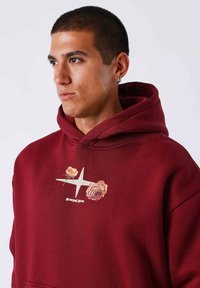 Sudadera burdeos con un gráfico floral en el pecho y un logo. Tejido suave, corte relajado y bolsillo tipo canguro. Capucha ajustable con cordón.